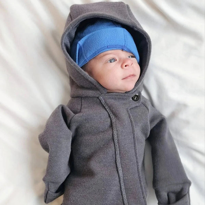 Baby in a gray hooded onesie and blue hat lying on a white surface