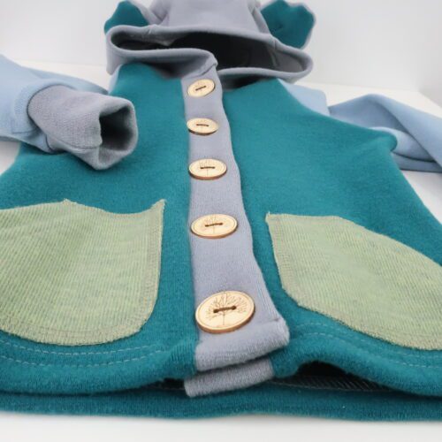 Merino Wool Button-Up Cardigan Sweater. This sweater has buttons the whole length of the sweater on a button panel that also runs the whole length of the sweater. There are options for a cuffed or hemmed bottom and sleeves are cuffed. Neck can be a short collar, tall collar, or a hood. This sweater is teal with light blue arms, a grey hood, button panel, and sleeve cuffs. There are additions of rounded angled pockets and bear ears on the hood. The buttons are a light wood with an outline of a tree on them.