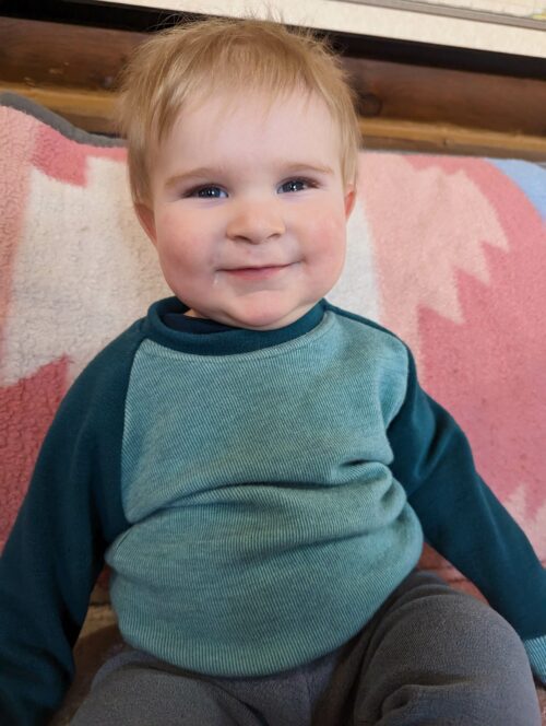 Merino Wool Simple Crew Neck Sweater. With cuffed raglan sleeves and a classic rounded crew neck. The child is wearing a striped medium green and dark green colour crew neck.